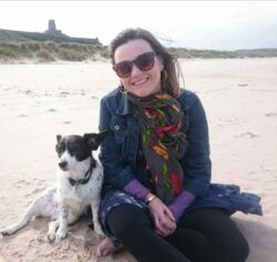 image description: Lisette, a white woman with freckles and brown hair, is sitting barefoot on a beach. She is smiling widely and is wearing sunglasses, a scarf, denim jacket and blue dress with leggings. Harper Lee, her black and white little rescue dog sits and leans beside her, her ears are sticking up because it’s windy. Bamburgh Castle sits behind them on the horizon.